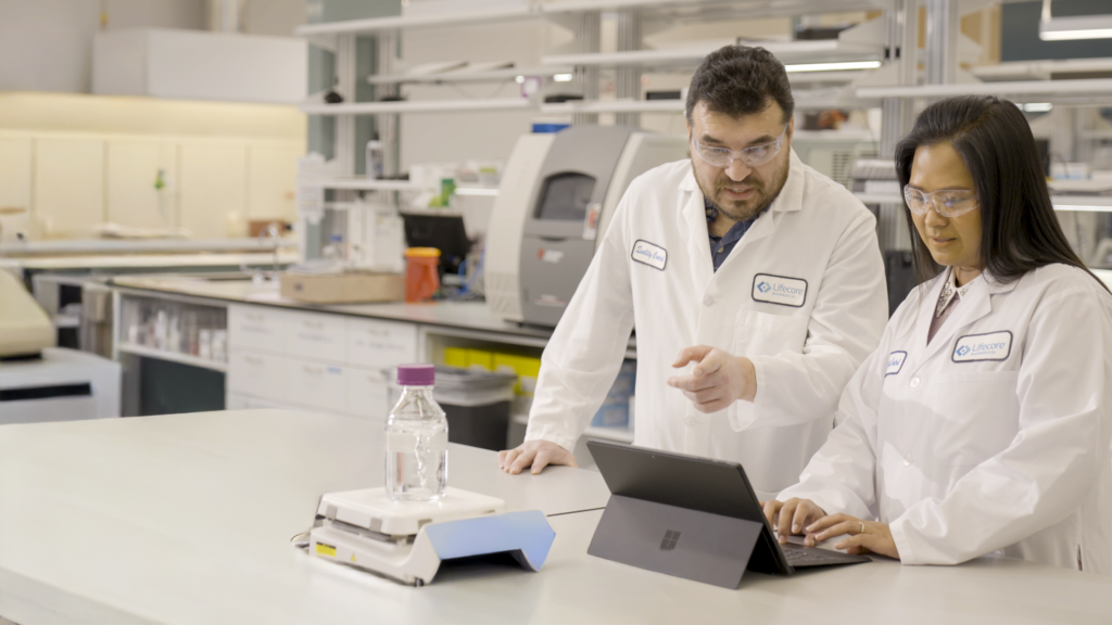 Male and female scientist in a lab, talking discussing data on a laptop.
