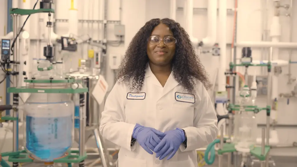 Lifecore Biomedical development team member in white lab coat and safety glasses, wearing purple nitrile gloves, standing in a biomedical manufacturing lab with stainless steel bioreactor equipment and glass vessels in the background.